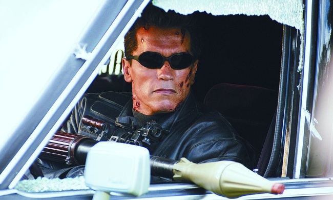 Arnold Schwarzenegger aims a rocket launcher out of a car window.